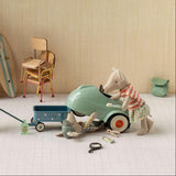 A styled miniature scene with small animal figurines and toy props arranged on a white floor against a beige wall. One figurine pushes a blue wagon while another stands beside a mint green toy car, with additional beach-themed accessories and furniture in the background.