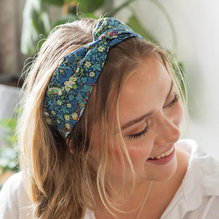 A smiling blonde woman wearing a blue floral knotted headband and a white collared shirt, photographed in a bright indoor setting with soft-focus green plants in the background.