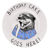 A white ceramic plate featuring a humorous illustration of an otter wearing a blue shirt with birthday cake slice patterns, surrounded by the text 'BIRTHDAY CAKE GOES HERE!'.