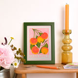 A framed art print featuring a vintage-style crab apple illustration in orange, yellow, and green on a pink background, styled on a wooden table with a pink flower, a candle holder, and a pencil.