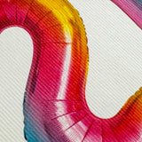 A close-up of a metallic foil number 2 balloon with a vibrant rainbow gradient transitioning from yellow and orange to pink, red, and blue, set against a white textured background.