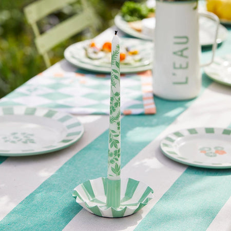 A tall white taper candle with a green herb leaf pattern stands in a matching green-and-white striped paper candle holder on a styled outdoor dining table. 