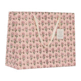 A pink paper gift bag with an all-over floral print, featuring cream-colored rope handles and a small hanging tag.