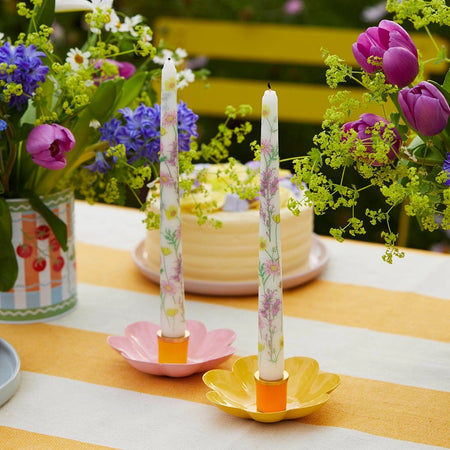 Decorative candles with floral patterns on a table with flowers and a cake in the background.