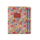 A floral notebook with a floral multicolor print of red, yellow, purple, and green flowers, featuring a brown leather-look label on the front that reads ' NOTEBOOK'.