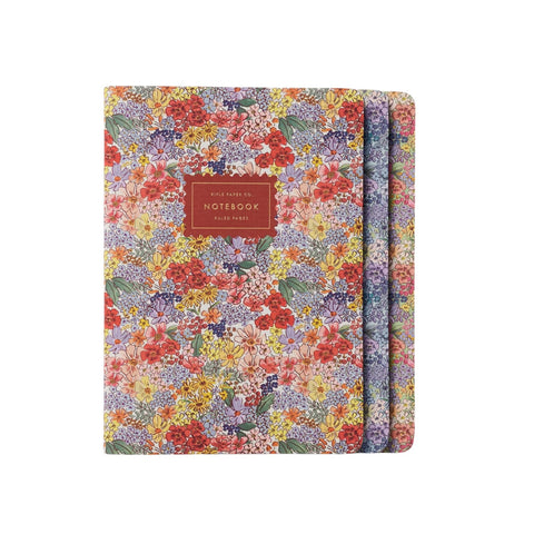 A floral notebook with a floral multicolor print of red, yellow, purple, and green flowers, featuring a brown leather-look label on the front that reads ' NOTEBOOK'.