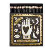 Matches box with decorative gold and white celestial symbols featuring a hand holding a heart on a black background with the text Miracle Luxury Matches.