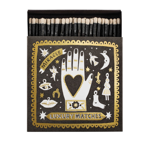 Matches box with decorative gold and white celestial symbols featuring a hand holding a heart on a black background with the text Miracle Luxury Matches.