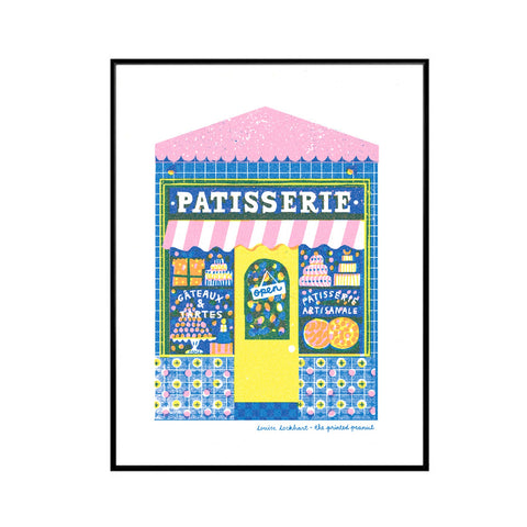 A framed art print featuring a colorful illustration of a French patisserie shop with a pink and white striped awning, blue patterned facade, yellow door, and various illustrated pastries. The print is presented on a white background inside a thin black frame.