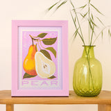 A framed art print featuring a pear illustration in autumn colors, styled on a wooden table next to a green glass vase with dried grass stems.