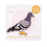 An embroidered iron-on patch of a pigeon, displayed on its original white backing card. The patch features a gray and purple body with black and white patterned wings and a bright yellow beak and foot.