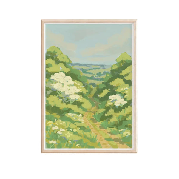 A framed landscape art print depicting a green valley path with white wildflowers under a pale blue sky, set in a light natural wood frame against a white background.