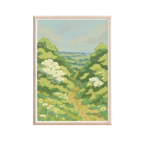 A framed landscape art print depicting a green valley path with white wildflowers under a pale blue sky, set in a light natural wood frame against a white background.