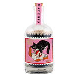 Matches in a glass bottle with a cork covered by a pink sticker with the word Matches in black. A decorative sticker covers the centre of the bottle with 1 black cat and 1 ginger and white cat playing with a Christmas ball ball on a pink background with Noel written in gold.